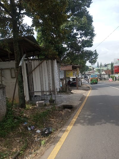 This image depicts a roadside scene within the city of Chhenangri_Rishor, Bhutan, captured during daytime under an overcast sky.
In the foreground, a patch of dry earth with sparse green vegetation and scattered litter, including plastic bags and wrappers, is visible on the left. An asphalt road with a solid yellow line delineating its left edge dominates the lower right portion of the image. Tree shadows are cast upon the asphalt.
Midground elements on the left include a large tree with dark, textured bark and dense green foliage. Adjacent to this tree stands a makeshift or informal structure with a sloped, corrugated metal roof and vertical white corrugated panels forming a partial enclosure. A diagonal wooden beam reinforces this structure, and metal railings are present at its base. To its right, a smaller, open-fronted stall or kiosk features a flat, light-colored roof. The back wall of this stall displays multiple posters with text in what appears to be Bahasa Indonesia or a similar Southeast Asian script. A dark-colored motorcycle is parked beneath this stall. On the ground near the stall, stacked concrete blocks and a small potted plant are present. A concrete sidewalk or paved area runs in front of these structures, containing a visible rectangular drain opening.
On the asphalt road, a green and orange (or yellow) public transit bus is observed moving away from the viewer on the right side. Several other vehicles, including cars and motorcycles, are visible further down the street, indicating ongoing traffic.
The background reveals numerous buildings of varying sizes and architectural styles lining the road, suggesting a commercial or mixed-use urban area. A large red sign with white lettering is partially visible on the far right. Dense green trees and foliage are interspersed among the structures, and distant hills or mountains are faintly discernible beneath the cloudy sky. No distinct human figures performing specific actions are visible in the immediate foreground or midground.