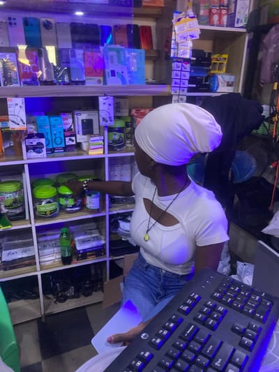 In Barakin, Nigeria, a young person wearing a white headscarf and a white cutout top with denim shorts is surrounded by shelves of electronics. The scene appears to be an electronics shop, with various items such as phones, chargers, headphones, and computer accessories neatly displayed. The person is looking down towards a keyboard and holding a white object, possibly a phone or a card, in their right hand. The lighting in the shop is dim, with blue and purple hues cast by the lighting, suggesting it might be evening or the lights are intentionally colored for ambiance. The shelves are filled with a variety of products, including boxes of phone accessories, power banks, and what appear to be phones themselves, some in packaging. The overall atmosphere suggests a busy retail environment, potentially with a focus on mobile devices and accessories. The text "14C", "Opay", "VGA", "TUNE TO", "Rm 14c", "FAST CO", "CABLE", "054", and "Iphors 11" are visible on various products and labels, providing clues about the types of items sold in the shop.