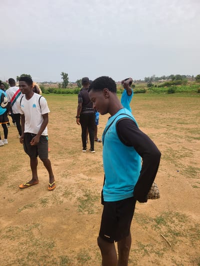 The image depicts a group of young men outdoors in what appears to be a field in Barikin Jirga Summa, Nigeria. The ground is a mix of dry dirt and sparse grass. The sky is overcast. The men are wearing a variety of casual athletic clothing. One man, positioned more prominently in the foreground, wears a white t-shirt, shorts and sandals. Further back, another man is wearing a blue t-shirt with black sleeves and shorts. The men seem to be stretching or engaged in a light warm-up activity. There are houses and some trees visible in the background, suggesting the edge of the city.