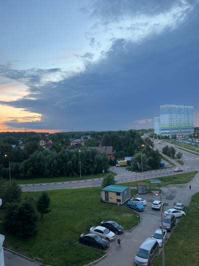 This is an aerial, eye-level, wide shot of a suburban landscape at dusk. A large, multi-story apartment building, with balconies and varying colored panels (white, light blue, green, and blue), dominates the right side of the frame. Adjacent to it is a smaller, similarly constructed building. A parking lot filled with various vehicles is situated in front of the apartment buildings. A road curves around the parking lot, with cars driving on it.
The foreground features a grassy area with several parked cars, including a dark sedan and a white car. A cyclist is riding a bicycle on a paved path. Further back, a dense line of trees separates the developed area from a collection of residential houses with red roofs. The sky is a gradient of colors, with a bright orange glow near the horizon on the left, transitioning to dark blue and grey clouds overhead. Power lines are visible in the distance. The overall scene suggests a residential area on the outskirts of Domodedovo, Russia, captured during twilight.