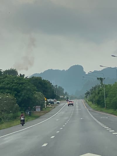 A photograph shows a curving asphalt road in Plai Phraya, Thailand. The road has white lane markings and white shoulder stripes. A red pickup truck and a motorcycle are on the road, heading in the same direction. Streetlights line the left side of the road. Trees and mountains are visible in the background. The sky is overcast. Some smoke is visible near the trees on the left.