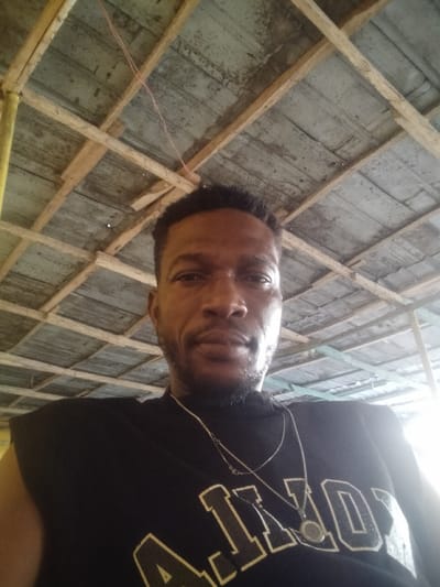A man with short hair and a trimmed beard poses for a selfie, wearing a black sleeveless shirt with white lettering and two silver necklaces, one with a pendant. The background reveals a ceiling made of wooden beams and corrugated metal sheets, with some light seeping in. The image suggests an informal, perhaps outdoor setting.