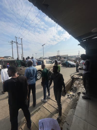 The image depicts a street scene in Akowonjo, Nigeria, with a group of people gathered around a pickup truck. The truck appears to be gold or tan colored and is filled with several individuals, some standing in the bed of the truck. Others are standing on the ground near the truck. The people are dressed in casual clothing. The environment appears to be an urban setting with buildings and utility poles visible in the background. The sky is partly cloudy. The foreground shows a sidewalk with some debris and a few individuals walking away from the camera. The overall impression is of a bustling street with a community gathering.
