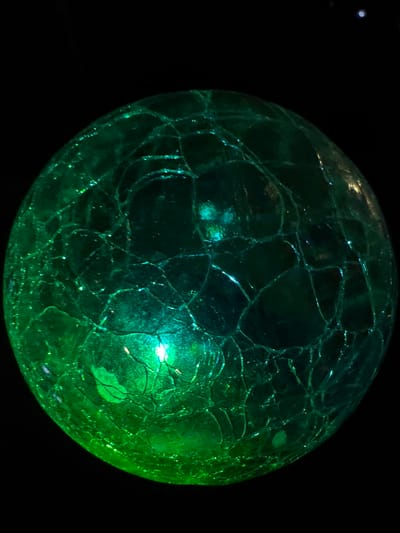This media file is a close-up photograph of a cracked, green glass sphere against a black background. The sphere is illuminated, with a bright light reflecting off its surface, highlighting the intricate network of cracks that cover its entirety. The cracks create a web-like pattern across the glass, giving it a textured and visually interesting appearance. The lighting causes some areas of the sphere to appear brighter and more translucent, while others are darker and more opaque. There are also small, sparkling highlights scattered across the surface, likely reflections from the light source. The overall impression is of a decorative object, possibly a marble or a bauble, displayed in low-light conditions. No people, text, or specific setting cues are visible in the image, other than the dark background which suggests an indoor or nocturnal environment. The photo does not convey any specific activity or event.