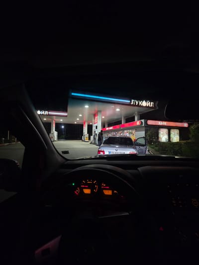 The image depicts a view from inside a car, likely at night, at a LUKOIL gas station. The gas station's sign is visible with the word "ЛУКОЙЛ" and a "КАФЕ" sign. A silver car is parked at one of the pumps. The speedometer and other dashboard elements of the car are partially visible, indicating the car is stationary. The location is identified as Momin Sbor, Bulgaria. A person appears to be near the pumps, presumably refueling the vehicle. The environment is illuminated by the gas station's lights.