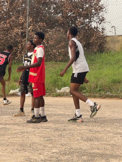 Four young men stand on a cracked concrete surface near a grassy area. One wears a white tank top with black shorts and white socks. Another has on a red and gold jersey with matching shorts and white socks. The third wears a black jersey with a graphic and dark shorts. The fourth person is wearing a black Jordan jersey with red number and gray shorts. The group appears to be a team, possibly after a game. A fence and some trees are in the background.
