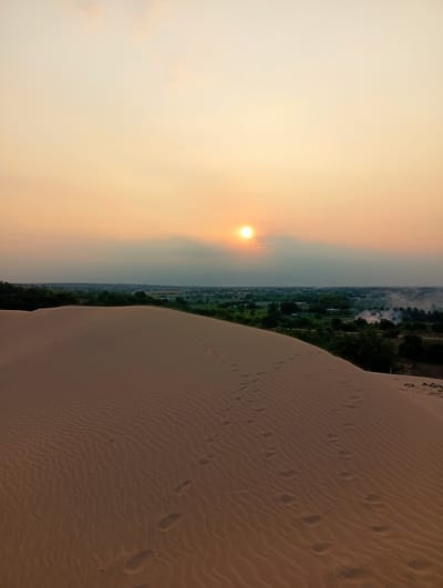 This image captures a vast desert landscape at either sunrise or sunset, specifically in Phuoc Dinh, Vietnam. The foreground is dominated by a large, undulating sand dune, rendered in warm, earthy tones. Its surface is intricately patterned with wind-blown ripples, and numerous human footprints are visible, trailing across the dune, indicating recent presence.
The sky above displays a soft gradient of orange and yellow, characteristic of twilight. The sun, a bright and luminous disc, is visible low on the horizon, partially obscured by light clouds or atmospheric haze, casting a warm glow across the scene.
Beyond the immediate sand dune, a strip of green vegetation stretches across the horizon, delineating the edge of the desert. Within this distant landscape, small buildings or settlements can be discerned, suggesting a nearby village or town. To the right of the distant settlement, plumes of white smoke are rising into the sky, hinting at some form of human activity, possibly burning. The overall scene evokes a sense of tranquility and the natural beauty of a desert environment at a golden hour. There is no visible text in the image.
