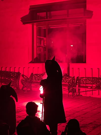 The image depicts a scene at night in Canet de Mar, Spain, illuminated by an intense red light. In the foreground, three figures are silhouetted against the light, two wearing dark, hooded cloaks resembling costumes, and one a young person facing away from the camera. To the left, another cloaked figure, possibly a performer, is seen holding a lantern. In the mid-ground, several ornate benches are visible, their dark metalwork casting strong shadows on the paved ground. Behind the benches, a building with a large window is present, revealing rows of bookshelves within. Wisps of smoke or fog are visible in front of the window, adding to the atmospheric and somewhat theatrical quality of the scene. The overall impression is of an outdoor event or performance taking place at dusk or night, with the red lighting creating a dramatic and slightly eerie ambiance.