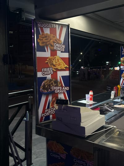 This media file shows a close-up of a food stall or kiosk, likely a fast-food vendor, at night. The primary focus is on a menu board displaying pictures of food items like onion rings, fried chips, chips and gravy, and a pizza portion, all set against a backdrop of the Union Jack flag. Some text visible on the menu board includes "NUGGETS," "ONION RINGS," "FRIED CHIPS," "CHIPS AND GRAVY," and "PIZZA PORTIO." To the right of the menu board, a cash register or payment terminal is visible, with "SAM4S" and "REGYSER" text on it. A small red and white bottle, possibly of sauce, is also present. In the background, through the glass or opening of the stall, a dimly lit street scene is visible, with blurred figures of people and building lights, suggesting an urban environment. The overall impression is of a busy evening service at a takeaway food establishment. The presence of "FIVE GUYS" and "FRESHER" text suggests this might be a local branch or a similar-themed establishment. The context of Benidorm, Spain, indicates a tourist-oriented location.