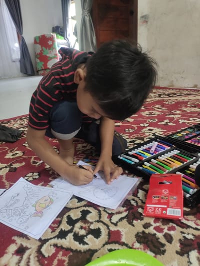 In a room in Cintadamai, Indonesia, a young boy, wearing a black and red striped t-shirt and dark shorts, is kneeling on a red patterned carpet and coloring in a coloring book. He is intently focused on the page, holding a black pencil in his right hand and a white pencil in his left. Beside him is an open coloring set with many colorful markers and crayons. The room appears to be a living area, with curtains visible in the background and a rolled-up mat with a floral pattern leaning against the wall. The lighting suggests it is daytime, with natural light coming from a window.