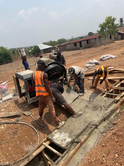 The image depicts an active construction site in Osara Gada, Nigeria, under a clear, bright sky. Seven male workers are engaged in concrete pouring operations on dry, reddish-brown earth. A large, black concrete mixer is centrally positioned. A worker in an orange safety vest and brown pants stands near the mixer, observing. Immediately to his right, a worker in dark clothing is bent over, emptying wet concrete from a large metal tray into an elongated wooden formwork trench. Another worker in a light-colored, striped shirt stands adjacent, possibly assisting with the concrete transfer. Further right, a worker wearing a yellow safety vest and brown pants kneels beside the partially filled formwork, actively shaping the freshly poured concrete. In the left background, a worker in a blue shirt carries a white bucket on his head. A worker in a dark cap and striped shirt is visible behind the concrete mixer. Multiple bags of cement are stacked on the ground to the right of the formwork, alongside wooden planks. A shovel and a black hose lie on the ground near the mixer. The background features several single-story buildings with corrugated metal roofs and sparse vegetation.
