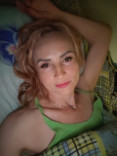 A woman with blonde, wavy hair and makeup lies on her back in bed, propped up on her left arm. She is wearing a green tank top and a silver necklace. The background consists of pillows and bedding with a checkered pattern. The image appears to be a selfie, taken from a slightly elevated angle. There is no information in the image to suggest a location in Shalsky, Russia.