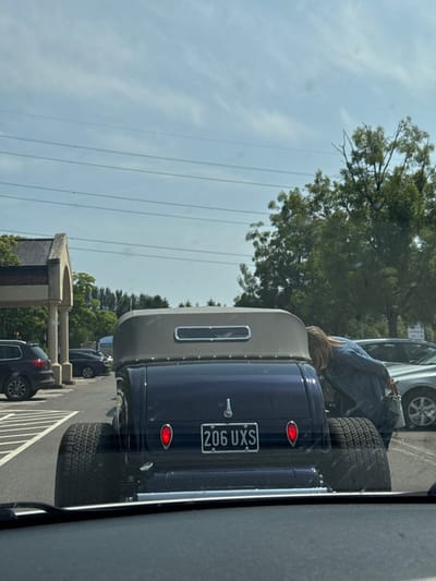 A blue vintage car with oversized rear tires is parked in a lot. The car has a light gray camper top and a license plate reading "206 UXS". A person with long blonde hair is leaning into the car's trunk. Other cars are parked in adjacent spaces. A building with an arched entrance is visible in the background. Overhead power lines stretch across the sky.