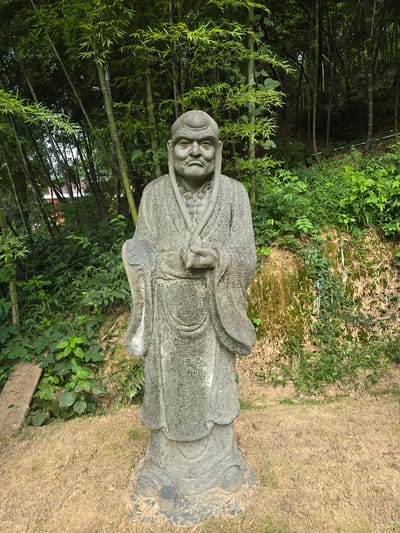 A stone statue of an elderly, bearded man stands in a garden. The man is wearing traditional robes and a cap, and he is holding his hands together in front of him. The statue is weathered and shows signs of age. The background is a dense bamboo forest, with lush green foliage and tall, slender bamboo stalks. The ground in front of the statue is covered in dry, yellow grass. The overall impression is one of peace and tranquility, with the statue appearing to be a guardian of the sacred grove. This statue is located in Xiaoshan District, China.