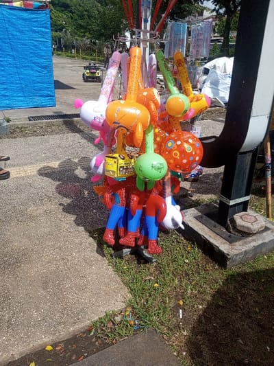 A colorful balloon stand displays an assortment of inflatable toys. Character shapes like Spiderman and cartoon animals are prominent. The stand is placed on a patch of grass next to a paved area. In the background, a blue tarp and a small electric car are visible. The scene is captured in daylight, creating a bright and lively atmosphere.
