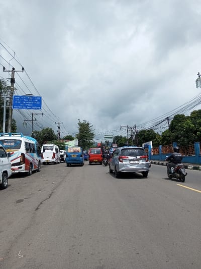 The image depicts a street scene in Indonesia, not Vaatharawattai, Sri Lanka, as indicated by the Indonesian text on the blue road sign visible on the left side of the frame.
The scene shows an asphalt road under an overcast sky, with vehicular traffic moving away from the viewer. On the far left, a white minivan with blue and red striped decals and "D'SALMA" text is parked or stopped, followed by two other white vans/minibuses. In the center lane of traffic, a blue minivan (license plate KT 1162 YI) with a rear window sticker is followed by a red minivan (license plate KT 1792 BN). In the right lane, a silver minivan (license plate KT 1215 YK) with a visible dent on its rear bumper is present. To the right of the silver minivan, a single individual wearing a black helmet and a dark jacket with "JC KILAU" or similar text is riding a black scooter-type motorcycle.
A blue rectangular road sign, mounted on a utility pole on the left, displays white Indonesian text listing locations such as "Terminal Tlp. 39 Minggu (24-01)", "Jl. Raya Nasional (24-01)", "S. Sidorejo (Dan Nasional) (24-01)", "Arah Kawasan", and "Pangkalan Loa Batas Gg. Jepong". Numerous utility poles with extensive, tangled overhead wiring are visible along both sides of the road. Green trees line both edges of the road, forming a background. On the right side, a blue painted concrete barrier with decorative patterns runs parallel to the road. The sky is uniformly gray and cloudy.