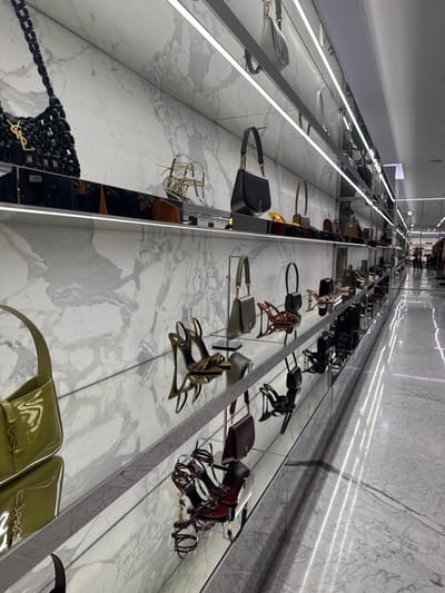 A luxury retail store, likely a Saint Laurent boutique, situated in Winchester, United States of America, is depicted from a slightly low-angle perspective looking down a long, brightly illuminated aisle. The walls are clad in white marble with grey veining. Merchandise is displayed on multiple levels of polished chrome or mirrored shelves, each featuring continuous strip LED lighting underneath. The floor is reflective, light-colored marble, mirroring overhead linear light sources. The primary subjects are high-end handbags and high-heeled shoes. Specific objects include a large black woven handbag with a gold 'YSL' logo, a black leather shoulder bag with a gold chain, an olive-green leather shoulder bag on a display stand, and a lime-green patent leather shoulder bag with a 'YSL' logo. Various high-heeled sandals are displayed, including gold strappy styles, olive-green open-toe heels, and dark red/burgundy elaborate strappy sandals. In the far distance at the end of the aisle, two indistinct human figures are discernible, one of whom appears to be wearing a red garment. No specific actions or interactions are occurring; the scene captures a static retail display.