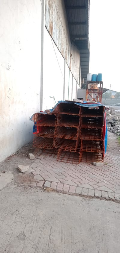 The image shows a stack of rusty, metal cages partially covered by a blue and red tarp. They sit on a brick-paved area adjacent to a white, weathered wall and a corrugated metal building. In the background are a metal water tower holding blue containers and other construction materials, including concrete blocks. The photo was taken in the city of Sumput, Indonesia. No people are visible.