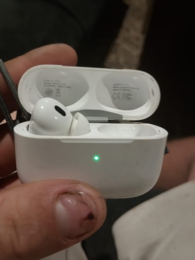 A hand holds an open white AirPods case. One AirPod is inside. A green light is illuminated on the front of the case. The surrounding environment is blurred and indistinct. The location is not provided in the image and cannot be inferred.