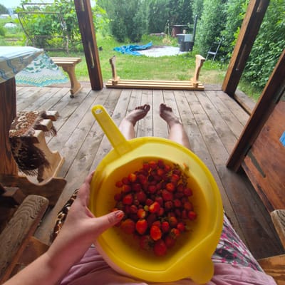 A first-person perspective shot shows a person sitting on a wooden deck in Novo-stroyka, Russia, holding a yellow colander filled with freshly picked strawberries. The person's legs, with bare feet visible, are extended forward. The deck has a wooden plank floor and is partially covered by a wooden roof supported by large wooden beams. To the left, a wooden bench with a patterned blanket is visible, and a table covered with a plastic sheet is also present. In the background, a grassy area features a swing set, some green foliage, and outdoor equipment including a grill or smoker and a blue tarp. The overall scene depicts a relaxed moment of harvesting and enjoying fresh produce in a rustic, outdoor setting.