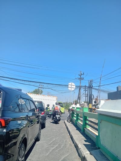 A vertical, eye-level outdoor shot captures a traffic jam under a clear blue sky in Gedangan, Indonesia. The foreground features the rear of a black car, its dark, reflective surface showing the sky and overhead wires. To its right, a row of vehicles and motorcycles are stopped on an asphalt road. Among them, a motorcyclist with a red backpack is visible. Further back, a white container truck is parked on the left side of the road. The background reveals numerous electrical wires strung across poles, a railway crossing signal, and a light green railing along a concrete barrier. The scene is illuminated by bright sunlight, casting shadows on the road and railing.