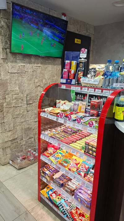 The image depicts the interior of a convenience store, likely in Nytva, Russia, judging by the Cyrillic text visible on the products. A large flat-screen television mounted on a stone-tiled wall displays a soccer match. To the right of the television, a door is marked with a yellow sign reading "B-4".

In the foreground, a refrigerated display case is filled with a variety of snacks, including candy bars, chips, and drinks. Several items are marked with "% АКЦИЯ" (meaning "SALE" or "PROMOTION" in Russian). Below the display case, on the floor, are several cans of Coca-Cola, some in a plastic bag, and a cardboard box. The store's flooring appears to be tiled. The overall lighting suggests an indoor environment, typical of a retail space. No people are visible in the frame.