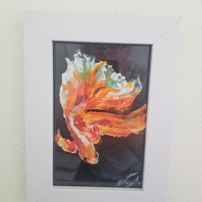 A framed painting of a fish is hung on a textured white wall. The painting features a vibrant, abstract depiction of a betta fish, rendered in bold strokes of orange, red, yellow, and hints of blue and white. The fish is shown in profile, its fins spread wide, giving it a dynamic and flowing appearance. The background is dark, which makes the brightly colored fish stand out prominently. The frame is simple and white, with clean lines, and it is positioned slightly off-center on the wall. The wall itself has a subtle, rough texture, suggesting it might be stucco or a similarly finished surface. The lighting in the image is soft, indicating it was likely taken indoors during the day, with natural light. There is no visible text in the image.