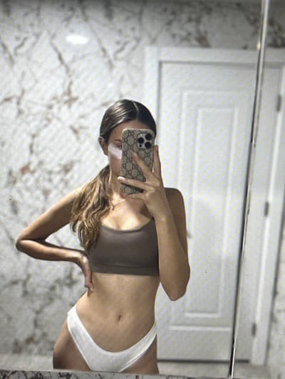 A young woman with her hair in a ponytail, wearing a brown sports bra and white underwear, is taking a mirror selfie in what appears to be a bathroom. She is holding a phone with a patterned case, angled to capture her reflection. The background shows a marble-patterned wall and a white door. There is no indication of the location being in Pruszcz Gdański, Poland.