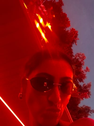 In a selfie taken at dusk in Sant Josep de sa Talaia, Spain, a person wearing sunglasses and an earring is illuminated by bright red lights, creating a dramatic effect against the twilight sky. The background features a tree silhouetted against the sky, with red lights also visible in the upper portion of the frame, likely part of an outdoor lighting installation or event. The overall atmosphere is vibrant and nocturnal, suggesting a nighttime outing or party.