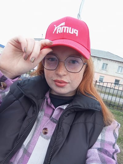 A young woman is captured in a close-up, slightly tilted portrait, seemingly a selfie, in Yurgamysh, Russia. She has reddish-auburn hair, fair skin with noticeable freckles, and light-colored eyes, which are visible through her thin-framed, hexagonal glasses. Her lips are painted a muted pink or coral shade.
She is dressed in a black zippered puffer jacket or vest worn over a plaid shirt featuring purple, white, and grey checks. A brown button is visible on her shirt. Underneath, a black top is peeking out. She is wearing a red baseball cap backwards, holding its brim with her right hand. Her fingernails are painted a muted taupe or grey color, each adorned with a thin, horizontal gold stripe.
The white text on the red cap, when viewed by the camera, reads "бМУЯ" in Cyrillic letters, appearing reversed and partially upside down due to the cap being worn backwards.
The background shows an outdoor setting with a light-colored brick building featuring several windows, likely residential or institutional. A metal fence or railing is visible between the subject and the building, with a patch of green grass at the bottom. The lighting is bright and diffused, indicating an overcast day during daylight hours. The overall impression is casual and relaxed.