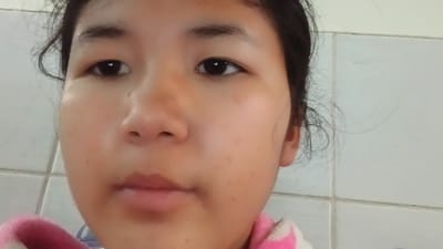 The media file is a close-up, head-and-shoulders portrait of a young East Asian girl. She is looking directly at the camera with a neutral expression. Her skin has a warm tone and appears to have some freckles or blemishes. She has dark hair, some of which is loose around her face, and her eyes are dark brown. She is wearing a pink and white garment, possibly a sweater or robe, around her shoulders. The background consists of white tiled walls, suggesting an indoor setting. The lighting is bright and appears to be from the front, likely from a flash or natural light source. There is no visible text in the image. The setting of Uchkun, Kyrgyzstan is a rural village in the Naryn region, known for its mountainous landscape. This image likely captures a moment of everyday life for the girl in her home environment.
