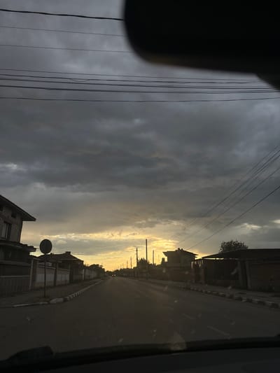 The image shows a street view in Manole, Bulgaria, at dusk. The sky is filled with dark, overcast clouds, with a strip of yellow light visible towards the horizon, indicating a setting sun. Utility lines are strung across the sky. The road stretches into the distance, lined with buildings on both sides. A road sign is visible on the left side of the road. The view is partially obstructed by a dark object on the left, possibly a part of the vehicle from which the image was taken. The overall mood is somber, reflecting the heavy clouds and low light.