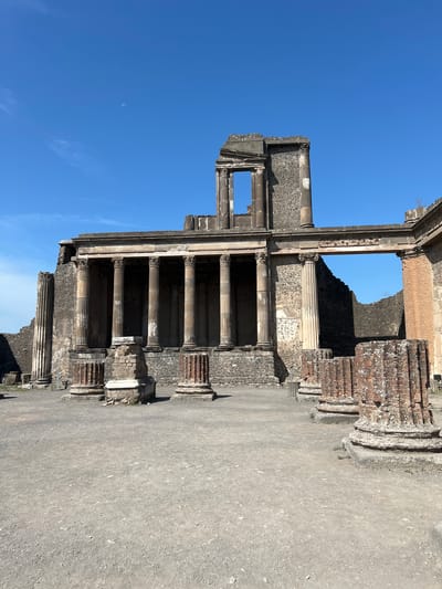 The image depicts the ruins of an ancient Roman building, likely a temple or public structure, at the archaeological site of Pompeii, Italy. The structure features a colonnade with Corinthian columns, some intact and others partially collapsed, made of stone and brick. Above the colonnade, a weathered entablature and pediment are visible, with a large rectangular opening in the upper section revealing the sky. The surrounding ground is a dusty, gravelly expanse, characteristic of excavation sites. To the left, a tall, fluted column stands, and to the right, fragmented columns and stonework are scattered. The sky is a clear, bright blue with a small, faint crescent moon visible. The overall scene conveys a sense of historical preservation and the passage of time.