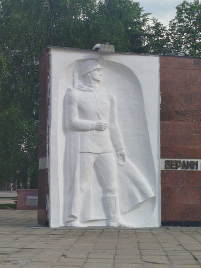 The image shows a Soviet War Memorial in Noginsk, Russia. The monument is made of red granite and white stucco. The white stucco part features a relief of a Soviet soldier in uniform. To the side is written "Berlin" in Russian. The memorial is located in a park setting with trees in the background and a brick tiled walkway in front.