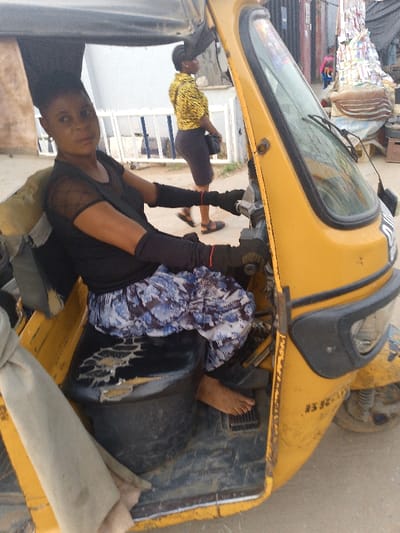 A woman sits in the driver's seat of a yellow three-wheeled auto-rickshaw, commonly known as a Keke Napep, in what appears to be a street or market setting in Ijaiye, Nigeria. She has short, dark hair and is wearing a black sheer-sleeved top over a black undershirt, black arm warmers, and a blue and white patterned skirt. She is barefoot, with her right foot resting on the vehicle's floor. Her hands are positioned on the handlebars or steering mechanism, and she looks directly towards the camera with a neutral expression.
The yellow vehicle shows signs of wear, particularly on the black driver's seat, which has visible tears and damage. The front of the vehicle, including the windshield and a single wiper, is partially visible on the right.
In the background, another woman is walking away from the camera, wearing a yellow and black patterned long-sleeved top and a dark skirt or dress. She carries a dark shoulder bag. Behind her, a white fence or railing is visible, and further to the right, piles of goods or market stalls suggest a bustling environment. The lighting is bright and diffuse, indicating daytime. Faint, indistinct text and numbers are visible on the lower front side of the yellow vehicle body.