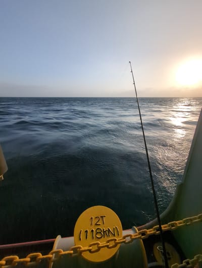 A fishing rod stands upright on a boat deck, casting its shadow onto the rippling, deep blue ocean. The sun, a brilliant orb of light, hangs low in the hazy sky, its rays reflecting off the water's surface in a dazzling display of light. A yellow-painted metal drum with the inscription "12T (118KN)" is visible in the foreground, wrapped in a yellow chain. The scene suggests a tranquil fishing excursion off the coast of Ussangoda, Sri Lanka, possibly at sunrise or sunset, given the low angle of the sun and the clear, expansive sky.