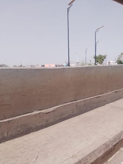 This image captures an urban streetscape in Chiranchi, Nigeria. The immediate foreground features a weathered, light-brown concrete barrier, approximately one meter high, spanning horizontally across the frame. Below this, a lower concrete surface is visible, forming a curb or sidewalk. Above the barrier, a series of dark-colored streetlights equipped with solar panels extend vertically against a hazy, light-gray sky. One prominent streetlight is positioned left of center, with several others receding into the distance on the right. In the background, distant low-rise structures with varying roof colors, including orange and white, are interspersed with scattered green vegetation, including a visible tree on the right. There are no individuals or discernible actions within the frame.