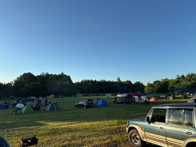 The image depicts a campsite set up in a grassy field on a clear day. Numerous tents of various colors, including blue, green, white, orange, and grey, are pitched across the landscape. Several vehicles are present, including motorcycles, cars, and vans, some of which appear to be parked near the tents. A prominent object in the foreground is a dark-colored grill. In the background, a dense line of trees forms the edge of the field. The scene suggests a recreational gathering or event. The location is identified as the Валожынскі сельскі Савет in Belarus.