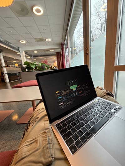 The scene depicts an individual seated in a public interior space, identified as a common area, likely within the Nord University, Levanger Campus, Norway. The perspective is from the seated individual's lap, looking slightly upwards and outwards.
The primary subject is a silver MacBook laptop, open and resting on the lap of an individual whose lower body is visible. The individual is wearing tan-colored cargo trousers with visible pockets and seam details, and their legs are crossed. The laptop screen displays a mind map graphic with a central node labeled "Weekly Schedule" and several branching nodes containing text and small icons. The MacBook's keyboard and trackpad are visible, featuring a standard QWERTY layout.
To the left of the laptop, a portion of a light-colored, slender-legged table is visible, beneath which pink, triangular floor panels are present on a speckled grey floor. An upholstered seat or cushion, colored orange-red, is partially visible under the individual's left leg.
The background shows a modern interior environment characterized by large, floor-to-ceiling windows with light wooden frames along the right side. Through these windows, bare trees with fine branches are visible against an overcast sky. The lower sections of some windows feature a frosted or opaque film. Inside, a spacious open-plan area extends into the distance, populated with various tables and seating arrangements. Several planters containing large green leafy plants and some plants with reddish foliage serve as dividers. The ceiling is a grid of white acoustic tiles, featuring multiple circular recessed LED lights with prominent rings and circular speaker grilles. Overhead, some areas in the deeper background display round, pendant-style light fixtures. The overall lighting is a combination of natural light from the windows and artificial interior illumination.