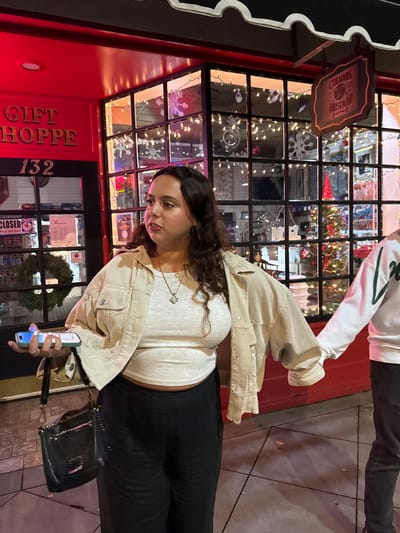 A woman stands in front of a store called "Gift Shoppe 132," illuminated with Christmas lights. She's wearing a beige jacket and black pants, engrossed in her phone. A person in a white sweatshirt walks by behind her. The storefront is adorned with holiday decorations.