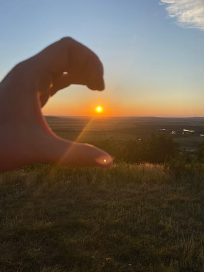 A human hand, silhouetted against the setting sun, is positioned in the foreground, obscuring a portion of the sky. The hand is curved, with fingers curled inward, creating a frame through which the sun is visible. The sun appears as a bright orange orb, low on the horizon. The sky transitions from a pale yellow near the horizon to a clear blue higher up, with a few wispy clouds present.
Beyond the hand and the sky, a vast, rolling landscape stretches out. This landscape consists of fields and scattered trees. Several bodies of water, possibly lakes or wide rivers, are visible in the mid-ground, reflecting the sunlight. The terrain appears to be slightly hilly. The foreground is dominated by tall, dry grass, illuminated by the warm light of the setting sun, creating a slightly blurred texture. The overall scene suggests an outdoor location at dusk, possibly a rural or natural setting. The specific location is Уязытамак, Russia.