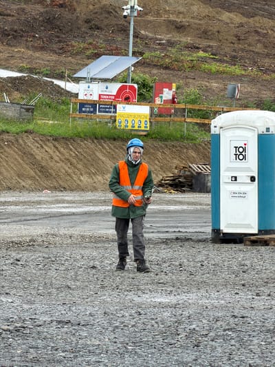 A construction worker, wearing a blue hard hat, green jacket, and orange safety vest, stands in the foreground of a construction site. He is looking towards the camera and holding a device in his right hand. Behind him, a portable toilet is visible on the right side of the image. The background shows a muddy embankment with some vegetation, solar panels, and various signs. The ground in the foreground is covered with gravel. This scene is likely located in Jodłownik, Poland, given the presence of Polish signage.