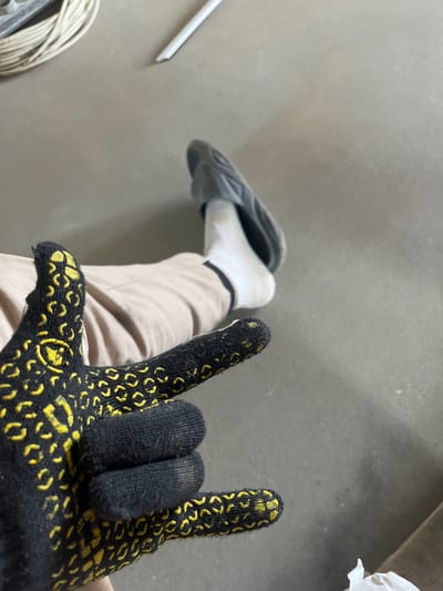A person wearing a black glove with yellow grip patterns is shown from the chest down, sitting on a gray floor in what appears to be an industrial or construction environment. The person is wearing light brown pants and white socks with gray slip-on shoes. The gloved hand is making a "rock on" hand gesture. In the background, a coil of white electrical wire is visible along with a white rod. The floor is concrete.