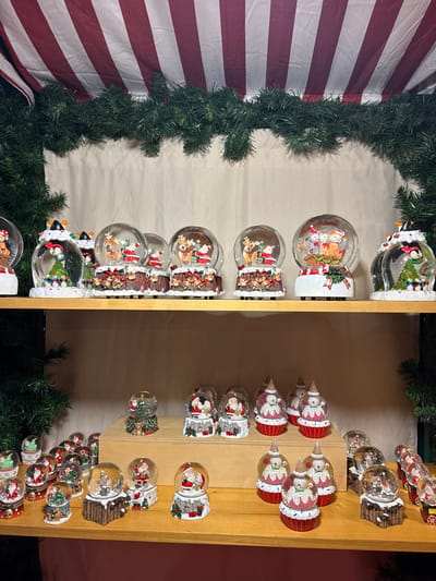 A shelf displays a collection of snow globes, mostly Christmas-themed. Many feature Santa Claus with reindeer, snowmen, or penguins inside. The bases are decorated in holiday styles, including festive red and white stripes, log cabins, and icy designs. A garland of green pine needles frames the left side of the shelf, while a striped red and white fabric hangs nearby.