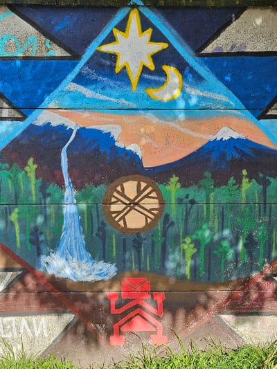 The image shows a section of a concrete wall adorned with colorful graffiti. The graffiti includes a blue diamond with a white star and crescent moon outlined in yellow, a peach-colored shape with white accents, a brown circle containing a stylized tree-like design, green and blue streaks resembling foliage and water, and a red geometric symbol possibly representing the letter "K". The concrete sections are separated by vertical lines. Some grass is visible along the right edge of the image. The location is Osorno, Chile.