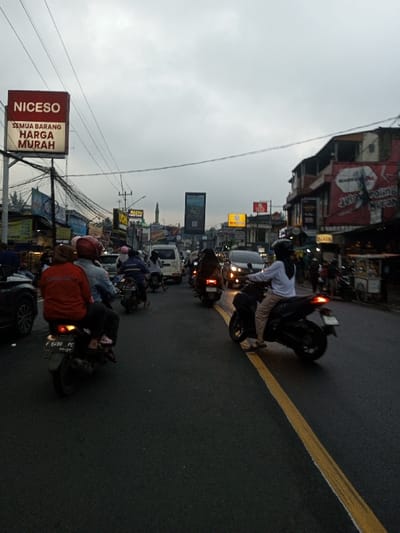 The image captures a busy urban street scene at dusk, likely in the city of Dawathang_Dorjibi_ Kashingtsawa, Bhutan, under an overcast, light gray sky. The asphalt road, appearing damp, features a prominent solid yellow line marking the center or a lane division. The scene is dominated by a dense flow of vehicles, primarily motorcycles, alongside cars and a white van, all moving away from the viewer.

In the foreground, on the left, the rear section of a dark car is partially visible. Behind it, a motorcycle carries two individuals; the driver wears a light blue jacket and a red helmet, while the passenger, positioned behind, wears a red jacket and a dark hijab. The visible license plate on this motorcycle is "F 8430 PC". In the foreground on the right, a dark scooter is ridden by a single individual wearing a white long-sleeved top, dark pants, and a dark hijab, with their left foot near the yellow lane marker. Numerous other motorcycles with riders, some wearing helmets and hijabs, are seen further down the road, interspersed with cars, including a dark sedan with illuminated headlights.

Both sides of the street are lined with multi-story commercial buildings featuring various shopfronts and prominent signage. On the left, a large red billboard displays "NICESO" at the top, with "SEMUA BARANG HARGA MURAH" (Indonesian for "ALL ITEMS CHEAP PRICE") underneath. An intricate network of overhead power lines and cables is visible above the street. On the right, other commercial signs are present, including one with text appearing to read "JELAJAH KULINER NUSANTARA" (Indonesian for "EXPLORE ARCHIPELAGO CULINARY"). In the mid-distance, a green dome and a minaret, characteristic of a mosque, are visible rising above the buildings on the left. The overall environment suggests a bustling commercial area with ongoing street activity.