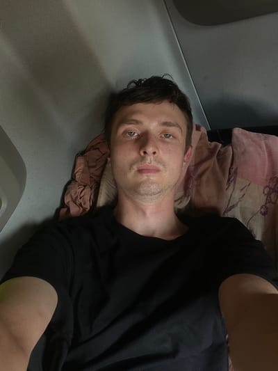 A man with light skin, short brown hair, and light green eyes is lying down on a surface that appears to be a bed or a mat. He is wearing a black t-shirt. A pink and beige patterned cloth is behind his head. The setting appears to be indoors, with a gray surface to his left, possibly part of a vehicle or furniture. The lighting suggests it might be nighttime or a dimly lit indoor environment. The man's expression is neutral, and he is looking directly at the camera. No text is visible in the image. The provided location context is Brachterbeek, Netherlands, but there are no visual cues in the image to confirm this.