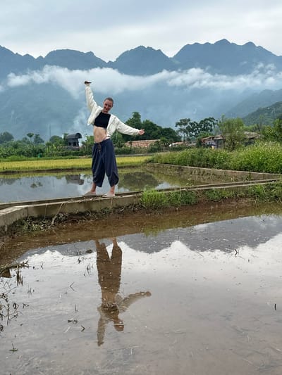 A young Caucasian woman with blonde hair pulled into a bun stands on a narrow concrete ledge dividing flooded rice paddies in Mai Chau, Vietnam. She is wearing a black crop top, a white jacket, and loose dark pants. Her arms are outstretched and her head is tilted back, as if in a moment of joy or celebration. In the background, lush green rice fields stretch towards mist-shrouded mountains. The water in the paddies reflects the sky and the woman, creating a mirror image. A few simple buildings and trees are visible in the mid-ground, adding to the rural atmosphere. The sky is overcast, with soft clouds hanging low over the mountains.