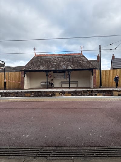 The image depicts the Benton Metro station platform in Newcastle upon Tyne, United Kingdom. The station features a covered waiting area with a tiled roof, ornate metalwork, and benches. A person is seated on a bench inside the shelter. Another individual stands further along the platform, towards the end of the platform structure. The platform surface is reddish-brown, with tactile paving along the edge. A section of wooden hoarding secured with red and white striped tape blocks part of the station structure. Overhead power lines are visible against a cloudy sky.