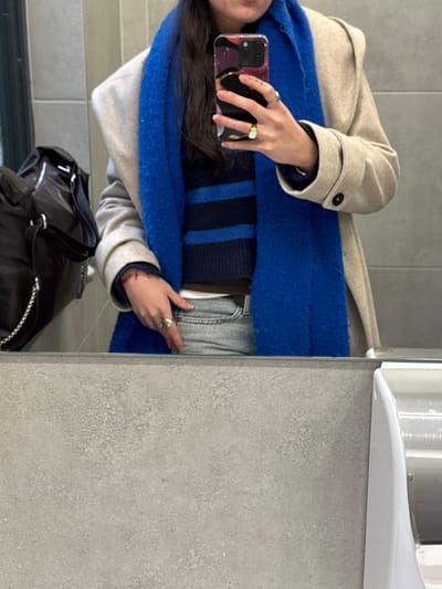 A person wearing a beige coat, a blue scarf, and a striped sweater takes a mirror selfie. The person is holding a phone with a colorful case. The reflection also shows a black backpack. The background consists of the mirror itself and a gray tiled wall with a white object mounted on it, potentially a dispenser. The overall composition highlights the clothing and accessories.